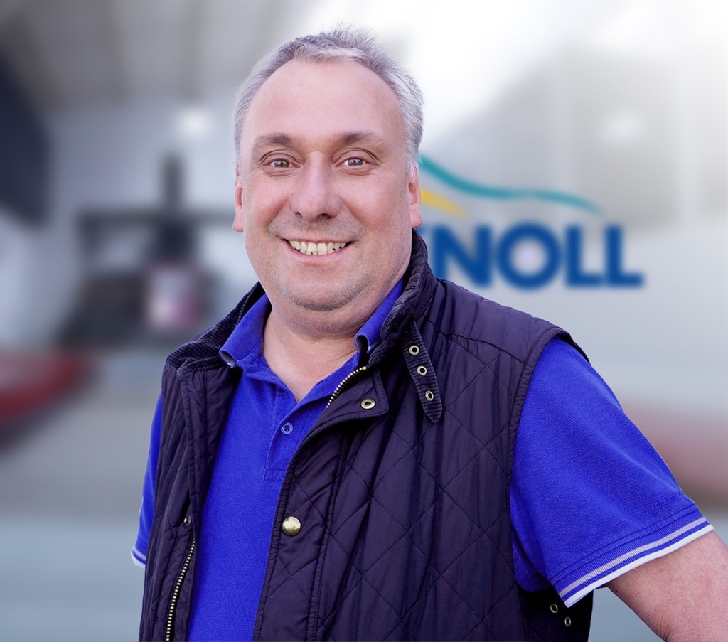 Ein lächelnder Mann mit kurzen, grauen Haaren trägt ein blaues Poloshirt und eine dunkelblaue Weste. Im Hintergrund ist das Wort "NOLL" sichtbar.