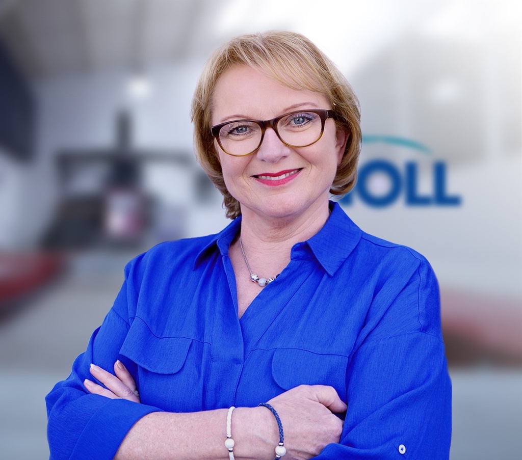 Eine lächelnde Frau mit blonden, schulterlangen Haaren trägt ein blaues Hemd und steht mit verschränkten Armen. Im Hintergrund ist unscharf das Logo eines Unternehmens zu sehen.
