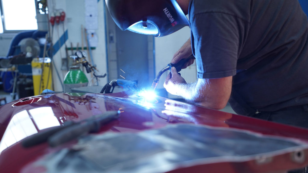 Ein Handwerker schweißt an einer glänzenden roten Metalloberfläche in einer Werkstatt. Funken fliegen beim Schweißen.