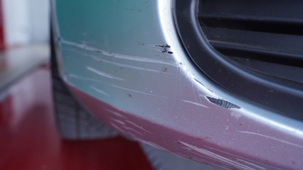 Der beschädigte Stoßfänger eines Autos zeigt mehrere Kratzer und Abplatzungen, die die silberne Lackierung beeinträchtigen.