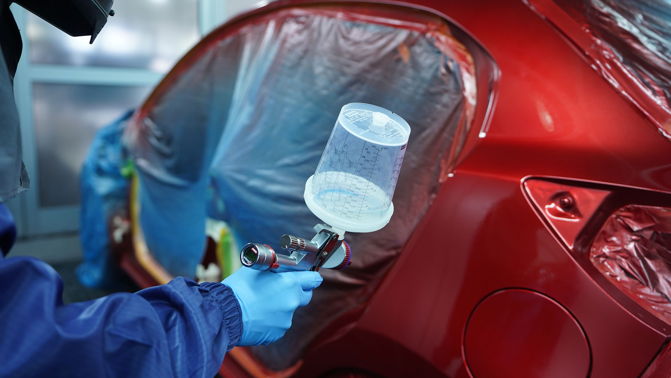 Ein Maler hält eine Spritzpistole in der Hand, während ein rotes Auto im Hintergrund lackiert wird.