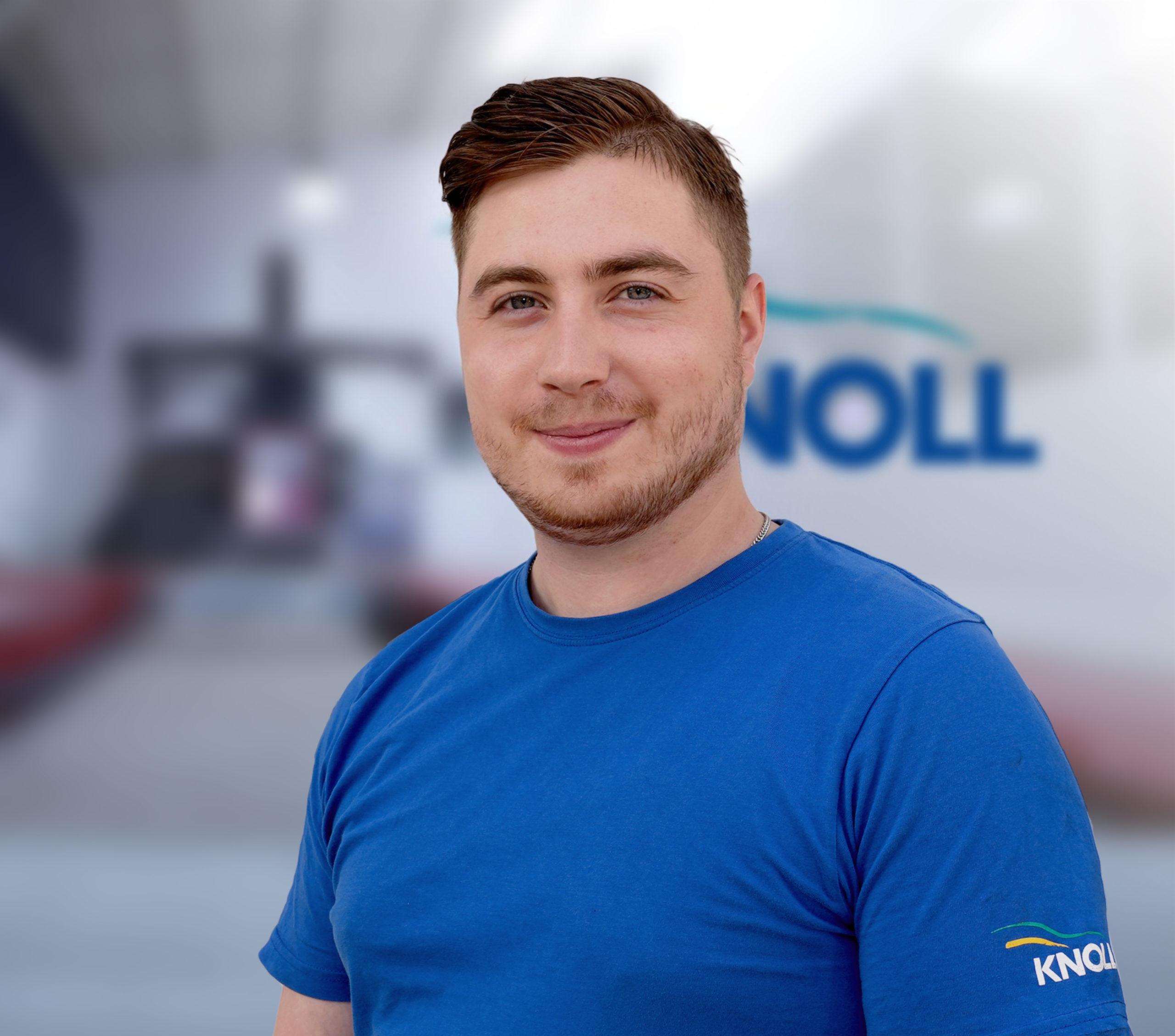 Ein junger Mann mit einem freundlichen Lächeln trägt ein blaues T-Shirt. Im Hintergrund ist verschwommen ein Firmenlogo zu erkennen.