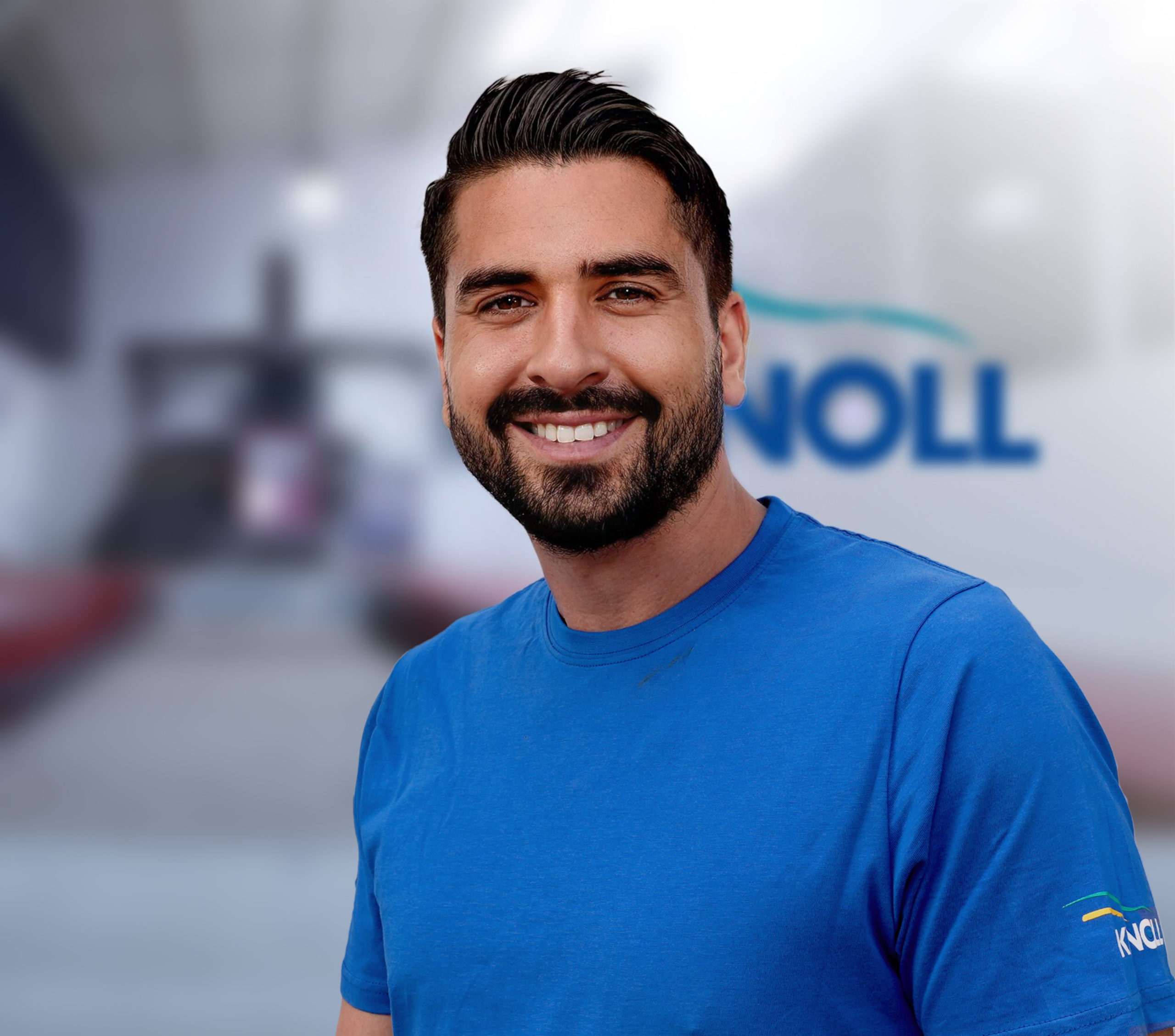 Ein lächelnder Mann mit dunklem Bart trägt ein blaues T-Shirt. Im Hintergrund ist unscharf das Logo "NOLL" zu sehen.