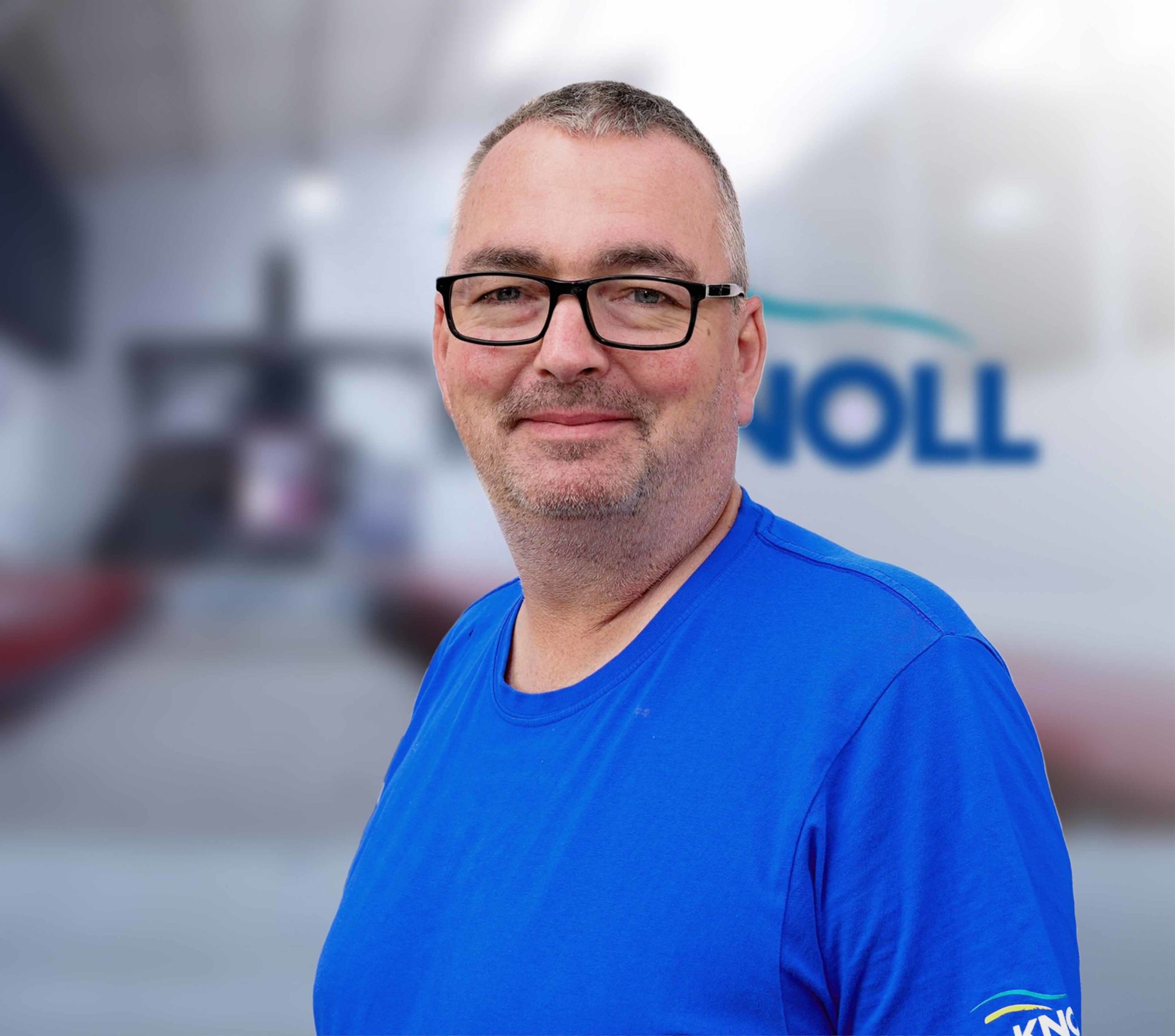 Ein Mann in einem blauen T-Shirt lächelt in die Kamera, mit verschwommenem Hintergrund und dem Schriftzug "NOLL".