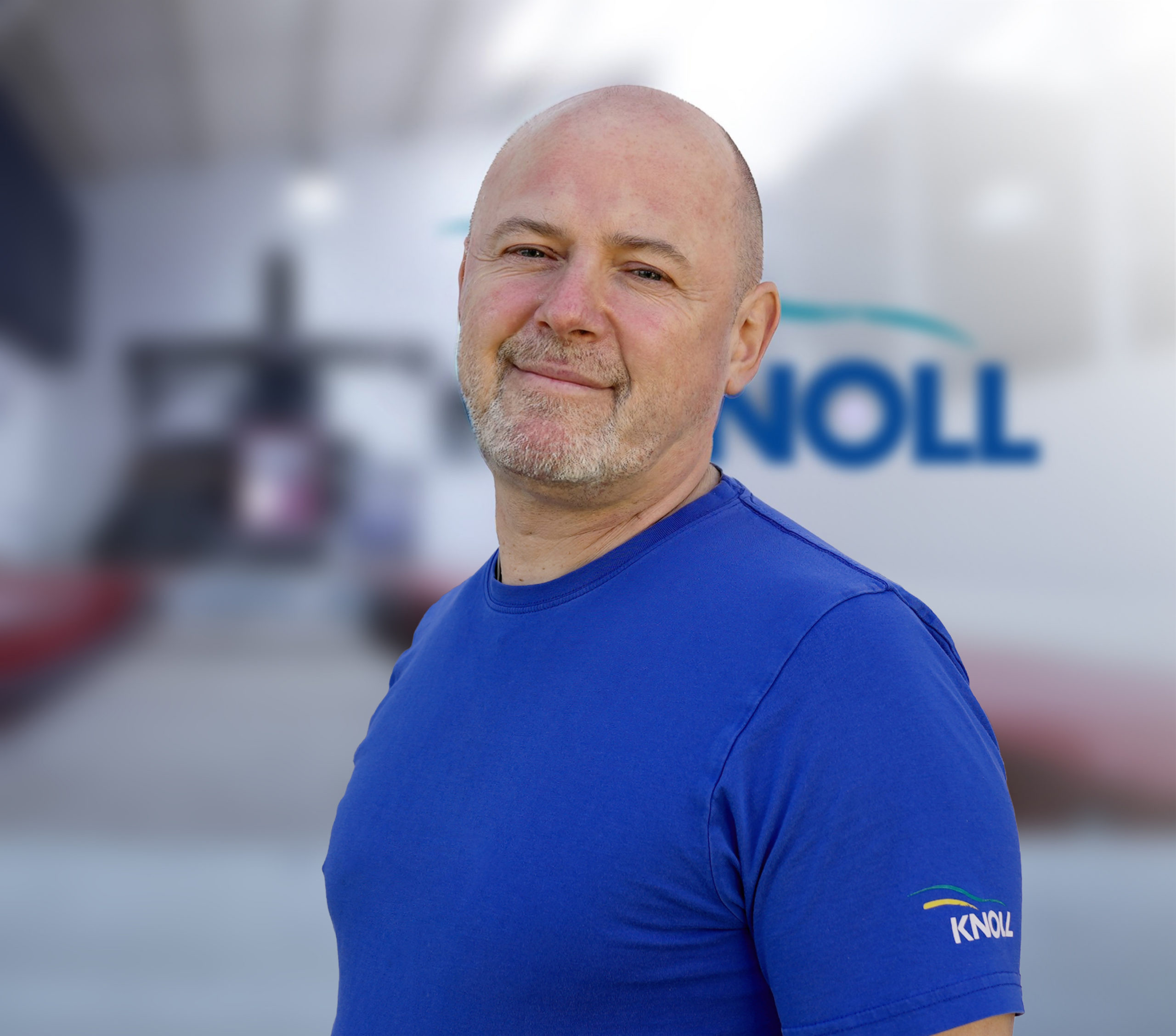 Ein Mann mit Glatze und Bart trägt ein blaues T-Shirt mit dem Logo "KNOLL" auf dem Ärmel. Der Hintergrund ist unscharf.