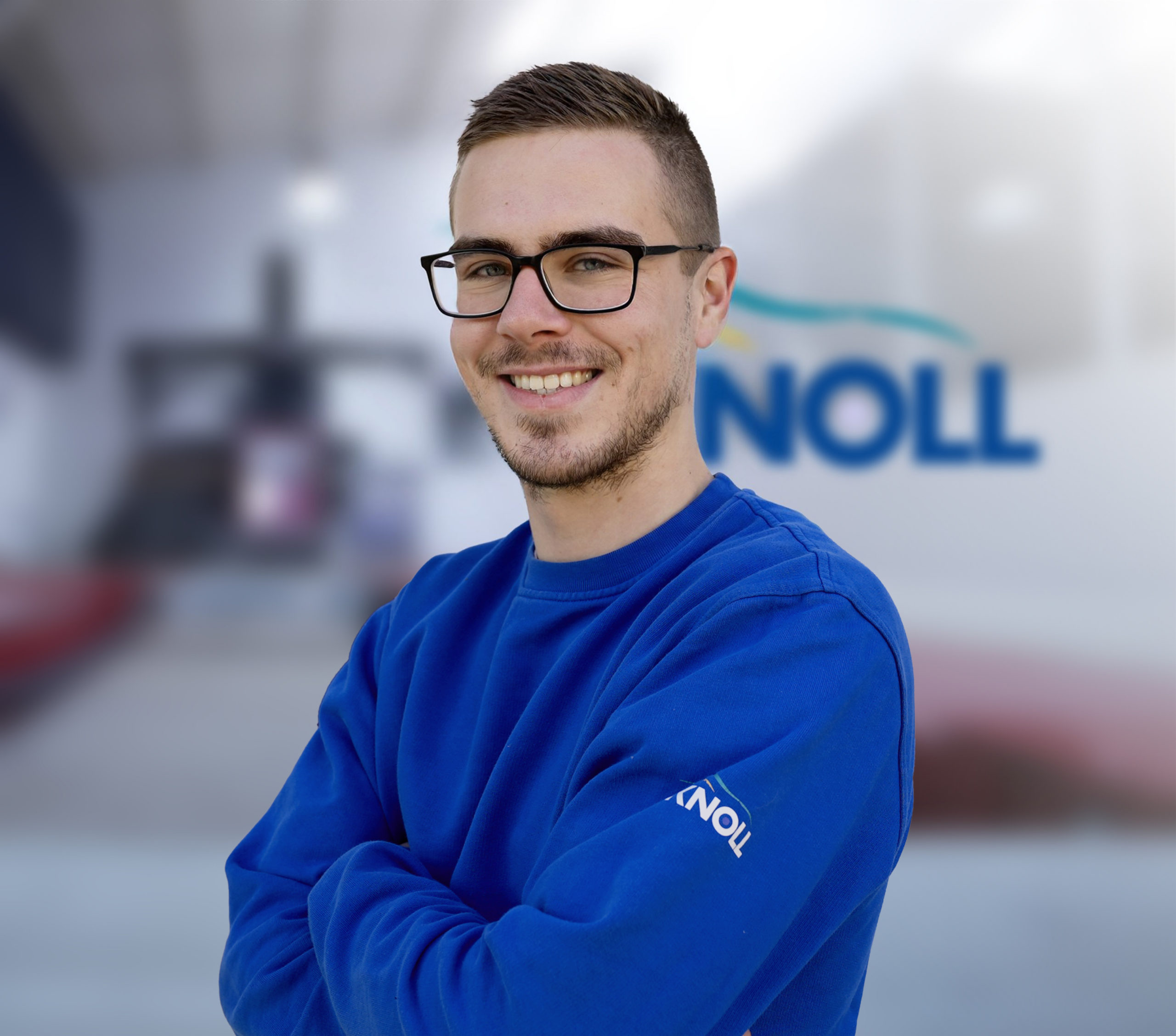 Junger Mann mit Brille und lächelndem Gesicht, trägt einen blauen Pullover mit Logo, Hintergrund unscharf.