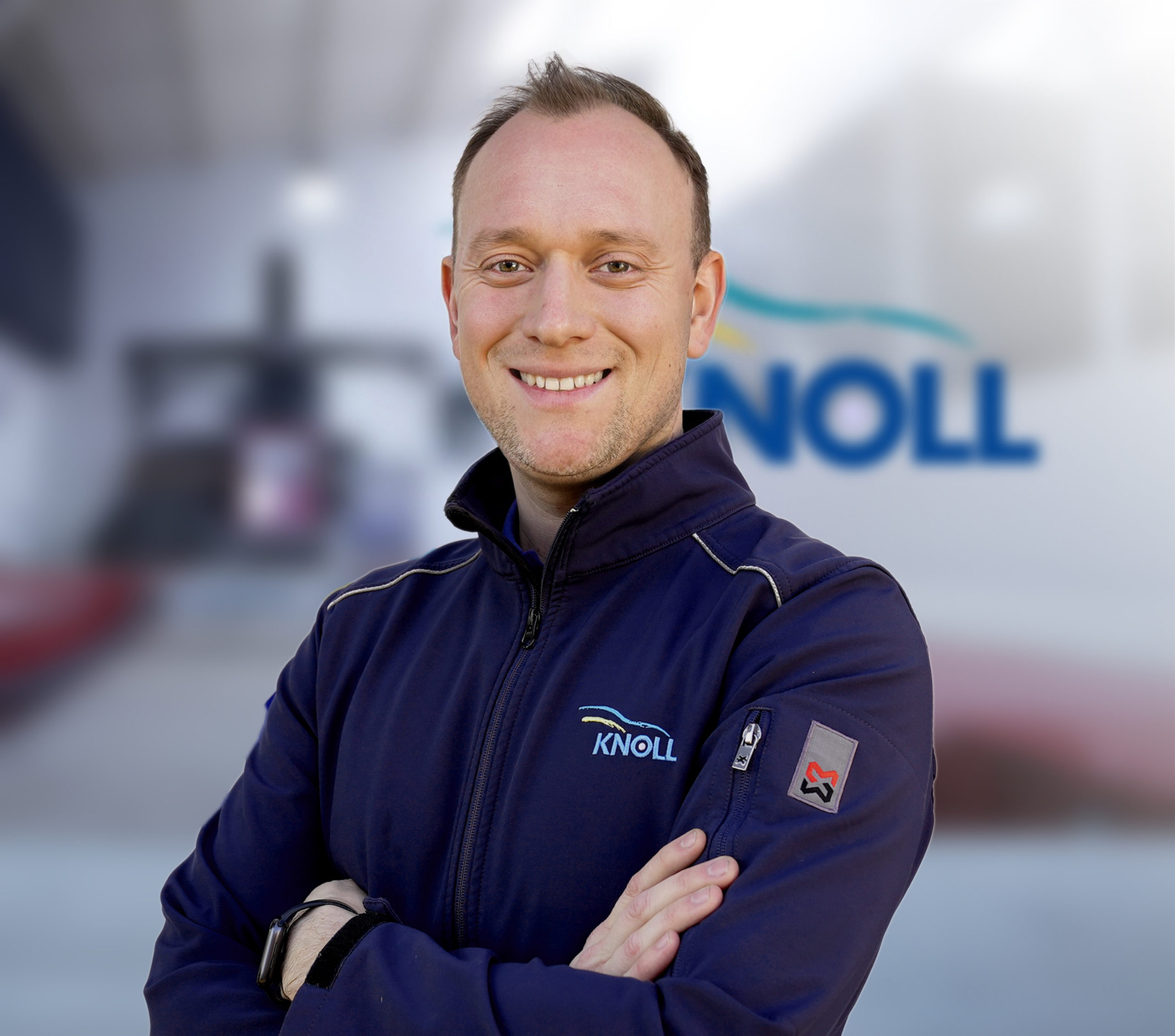 Ein lächelnder Mann in einer blauen Jacke mit dem Logo "KNOLL" steht mit verschränkten Armen vor einer unscharfen Hintergrundszene.