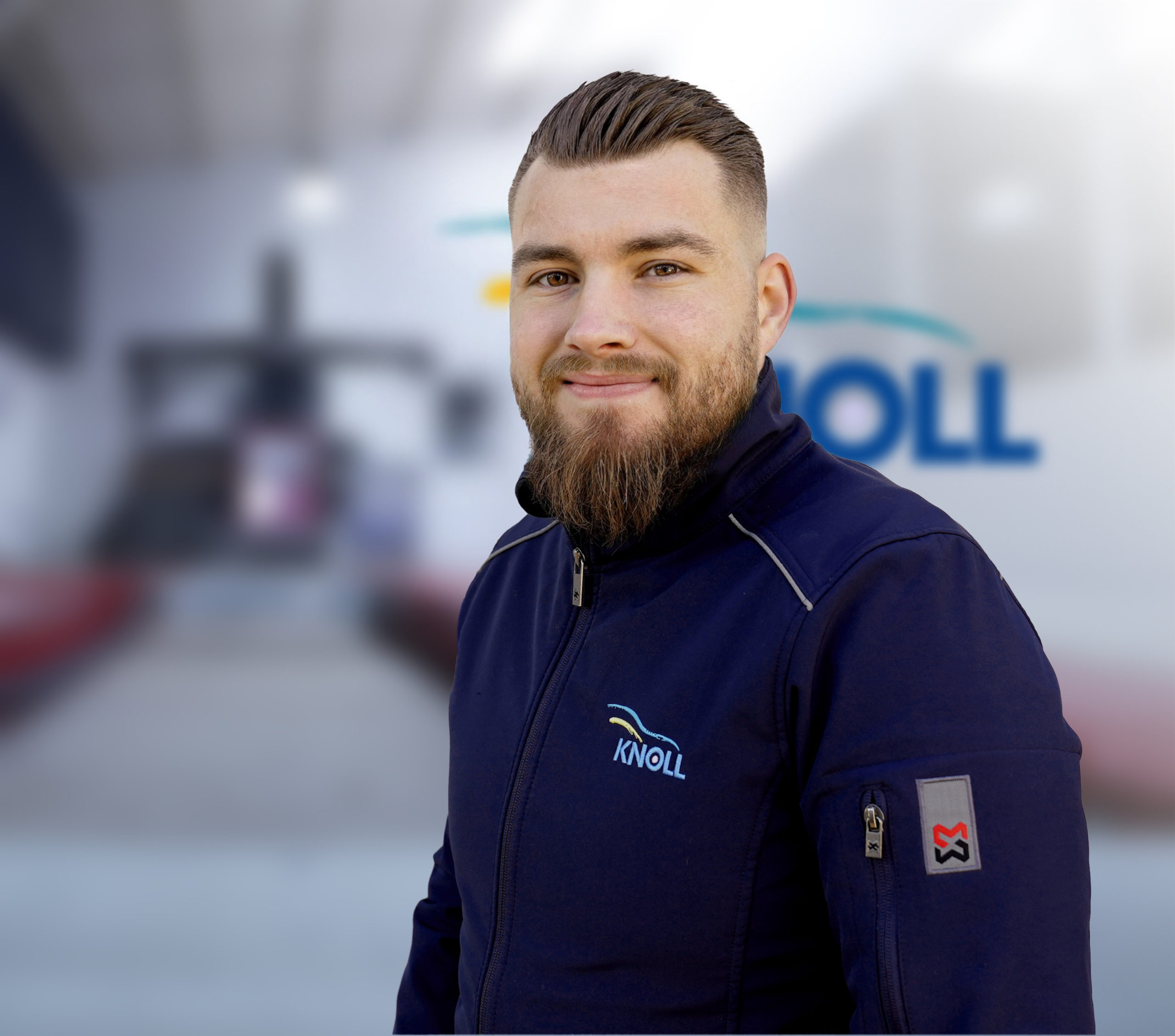 Mann mit brauner Bart und gestyltem Haar trägt eine blaue Jacke mit dem Logo „KNOLL“ in einer Werkstatt.