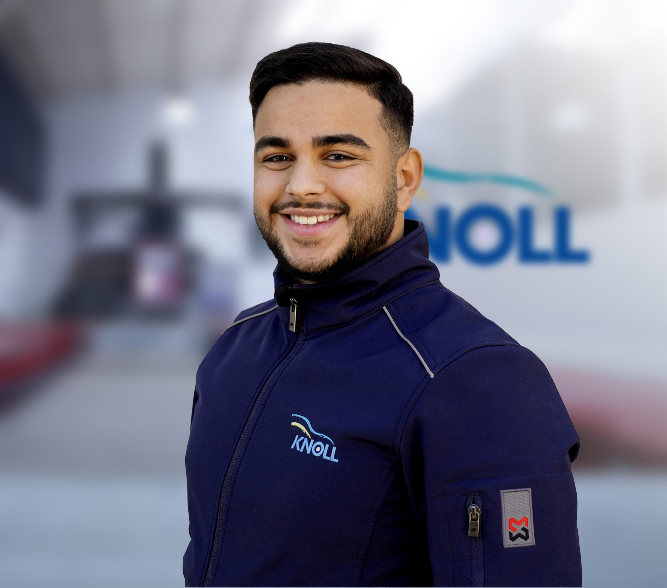 Ein lächelnder Mann trägt eine dunkelblaue Jacke mit dem Logo "KNOLL". Im Hintergrund ist eine unscharfe Werkstattumgebung zu sehen.