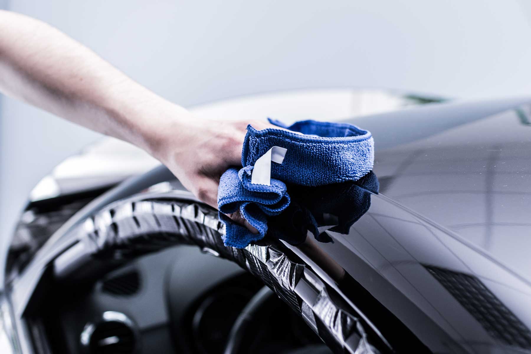 Eine Hand reinigt mit einem blauen Mikrofasertuch die glänzende Oberfläche eines Autos, während der Innenraum sichtbar ist.