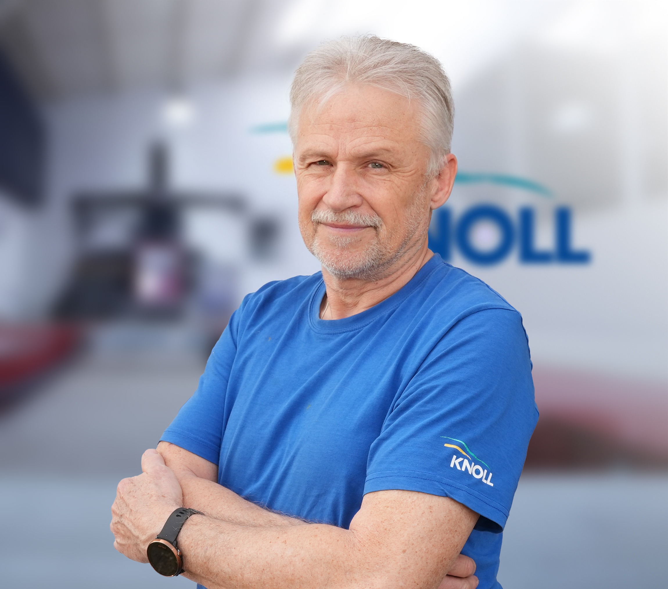 Mann mit grauen Haaren in blauem T-Shirt, lächelt und steht mit verschränkten Armen vor unscharfem Hintergrund mit Schriftzug.