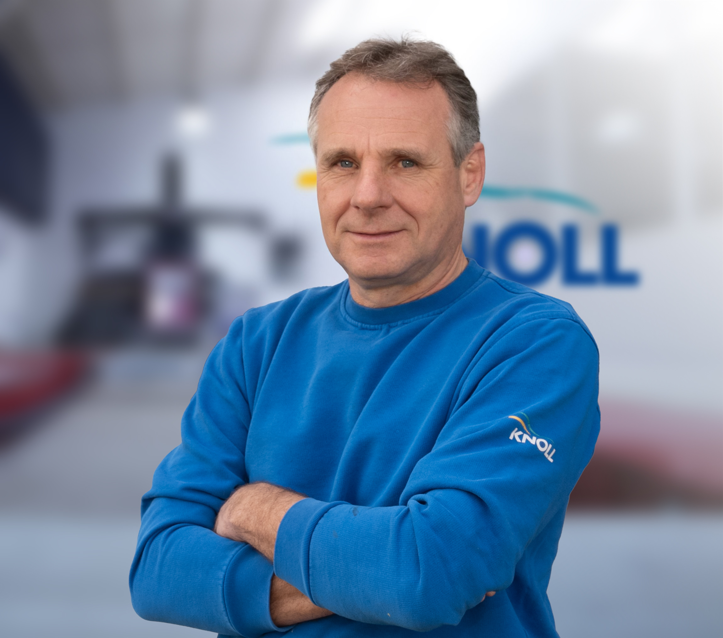 Ein Mann mit kurzen grauen Haaren trägt einen blauen Pullover und steht mit verschränkten Armen vor einem unscharfen Hintergrund.