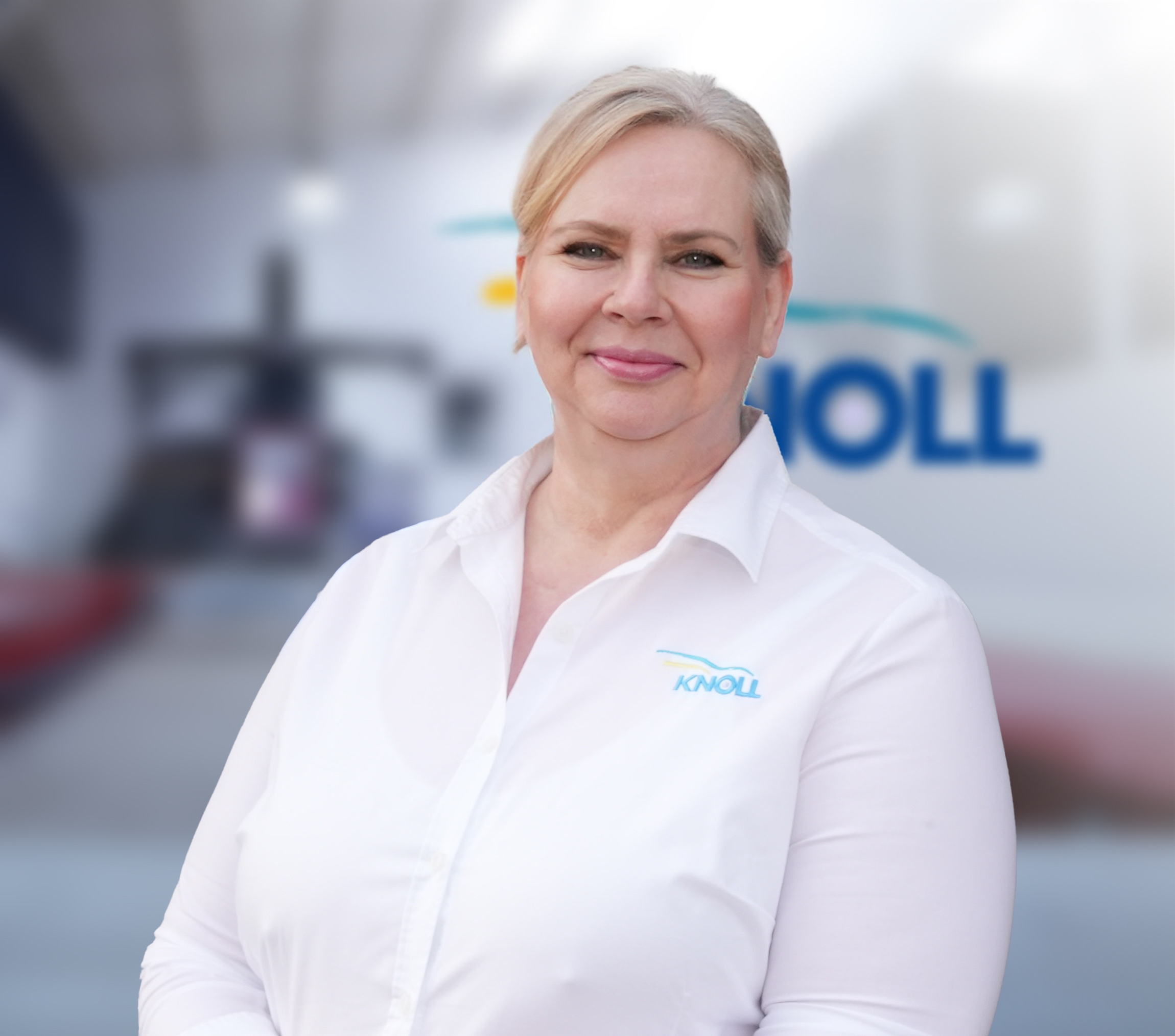 Eine Frau mit blonden Haaren trägt ein weißes Hemd mit dem Logo "KNOLL". Im Hintergrund sind unscharfe Maschinen und das Firmenlogo zu sehen.