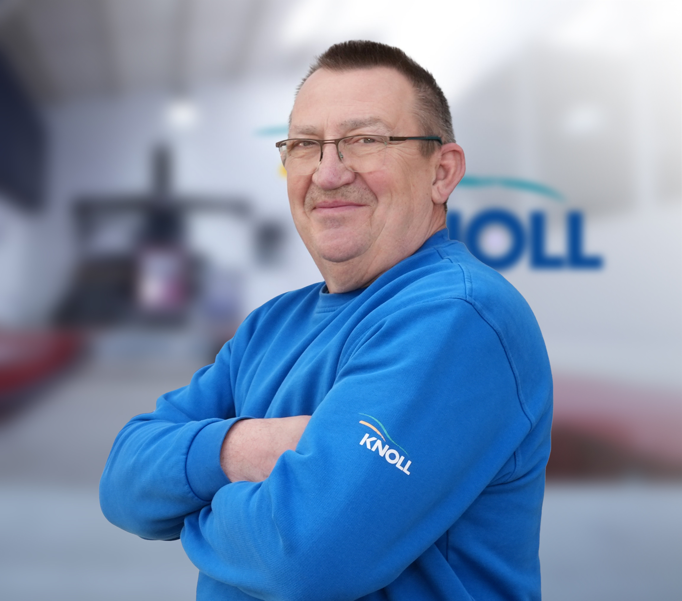 Ein lächelnder Mann in einem blauen Sweatshirt mit dem Logo "KNOLL" steht mit verschränkten Armen vor einem unscharfen Hintergrund.