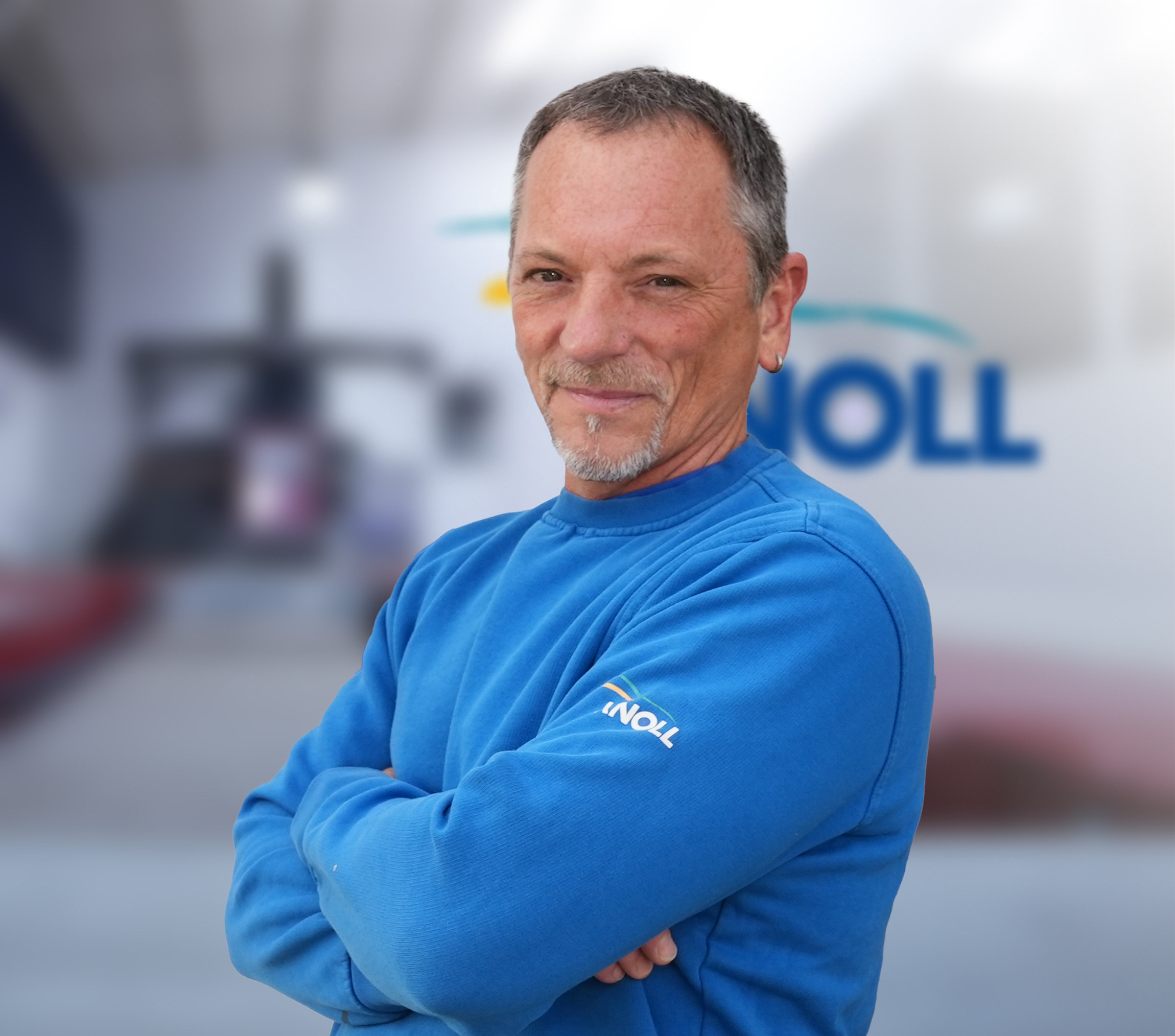 Ein lächelnder Mann in einem blauen Sweatshirt steht mit verschränkten Armen vor einem unscharfen Hintergrund, auf dem das Logo eines Unternehmens zu sehen ist.